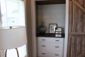 Built-in shelves with decor, drawers below, next to a window with blinds and a floor lamp in this 2 Beds swayback design.