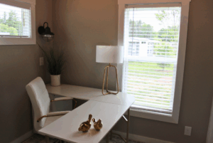 Small, modern home office with a white desk, chair, lamp, plant, and Swayback gold bird decor by a window.