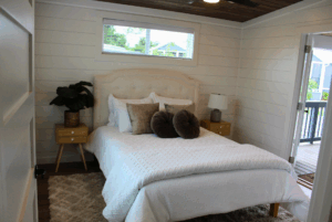 A cozy bedroom with a swayback beige headboard, white bed, pillows, nightstands, and a window showing greenery.