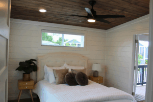 Cozy bedroom with wooden ceiling, white walls, 2 beds, nightstands, lamp, plant, and open door to a porch.
