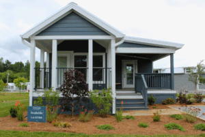 A small modern gray swayback house with 2 beds, front porch, garden, and an Available sign on the lawn.