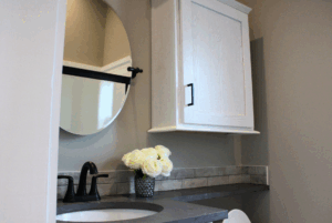 Modern bathroom with round mirror, black faucet, white cabinet, and 2 beds nearby for added comfort.