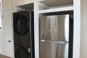 A stacked black washer and dryer swayback next to a stainless steel fridge in a white alcove.