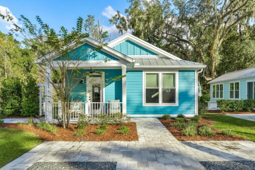 A small, blue, single-story house with white trim, and a front porch, situated in a garden with paved pathways. The house is surrounded by trees and greenery under a blue sky.