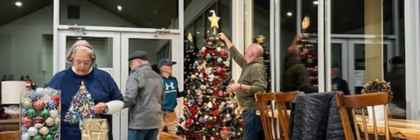 People decorating Christmas trees inside a room with large windows. One person stands with a small decorated tree, while others are placing ornaments and a star topper on larger trees.
