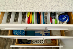 Aerial view of two open kitchen drawers: the top drawer contains organized utensils and kitchen tools; the bottom drawer holds various items including a tin star mold, cloth items, and a sealed gift box.