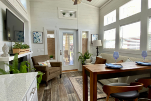 A cozy living room with white walls, a wood floor, a brown chair, a beige sofa, a tall wooden table with two stools, and large windows allowing natural light to fill the space.