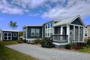 A small modern blue house on Strolling Easy Lane with a porch and shed, set beneath a partly cloudy sky.