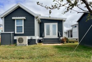 A small dark gray house with white trim sits on Strolling Easy Lane, steps leading to double doors and a grassy yard in front.