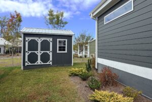 A gray shed with white trim sits on Strolling Easy Lane beside a gray house on a clear, sunny day.