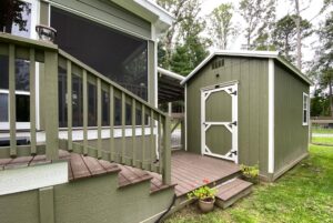 Green shed with white trim next to a porch with stairs at 108 Meandering Lane, surrounded by grass and potted plants.