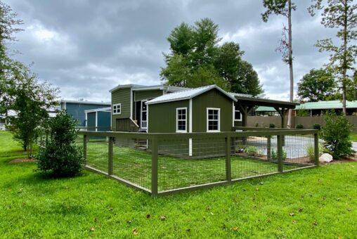 Charming green tiny house at 108 Meandering Lane with a fenced yard, nestled among trees and grass under a cloudy sky.