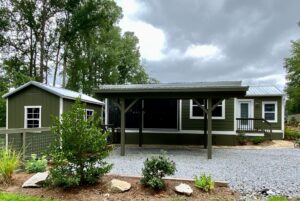 Charming green tiny house at 108 Meandering Lane with a covered porch, gravel drive, shed, and lush landscaping.