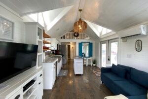 Bright, modern tiny house interior at 40 Drifting Brook Lane with white walls, kitchen, blue sofa, and pitched ceiling.