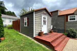 Small gray shed at 40 Drifting Brook with white door, red steps, and flower pots on a green lawn under a cloudy sky.