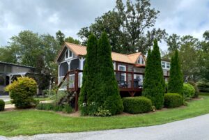 A small house with a porch on Drifting Brook Lane, surrounded by trees and manicured bushes on a cloudy day.