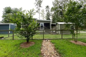 A small gray house with a porch sits on Meandering Lane, with a stone path, trees, and a wire fence in a green yard.