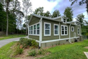 Green tiny house with white trim at 108 Meandering Lane, many windows, flower bed in front, surrounded by trees.