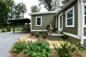 Green house with white trim on Meandering Lane, small porch, garden, and a gravel driveway with covered carport.