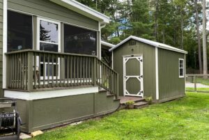 A green shed with white trim sits by a porch on the grassy lawn at 108 Meandering Lane, framed by trees in the background.