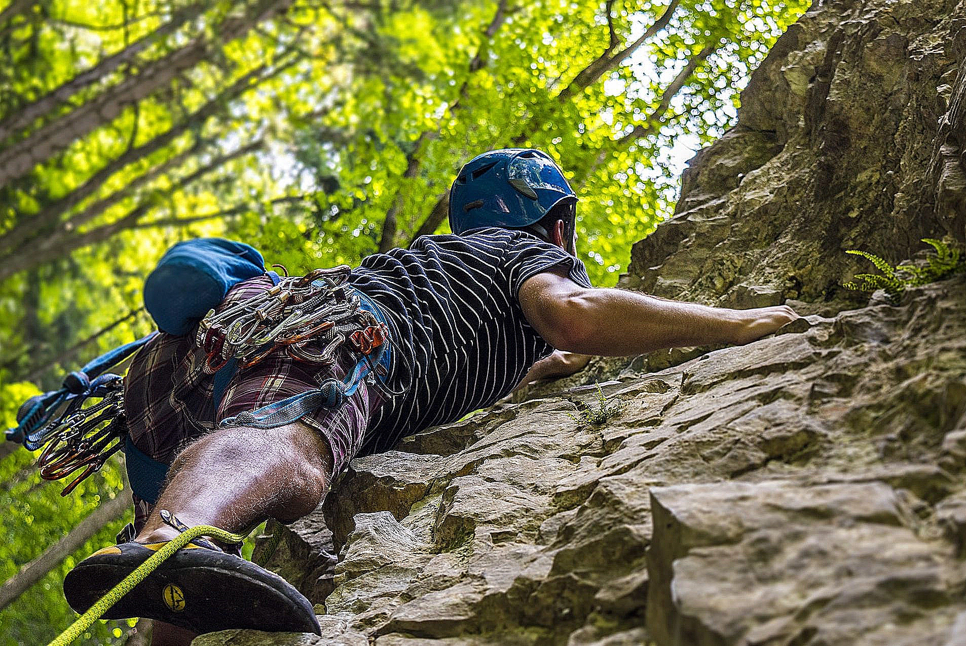 Person rock climbing outdoors at Flat Rock, NC, wearing safety gear and helmet, surrounded by trees and sunlight.
