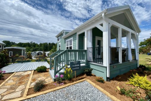 A small green house with a covered porch, surrounded by gardens, under a partly cloudy sky.
