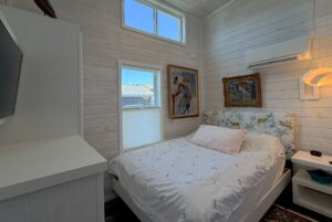 Small, bright bedroom on Skipping Stone Lane with white walls, bed, TV, artwork, and two sunlit windows.