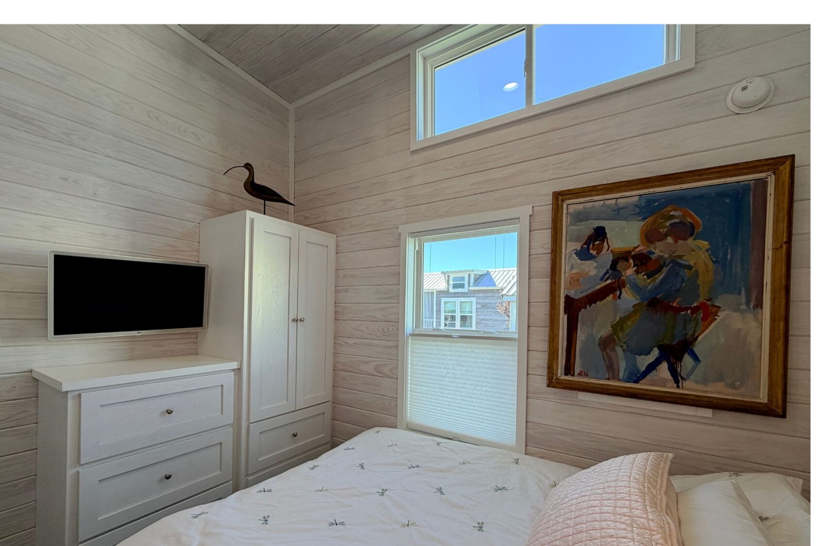 A cozy bedroom at 49 Skipping Stone Lane with white furniture, a TV, wall art, and sunlight through two windows.
