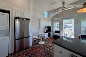 A bright, modern tiny home interior at 49 Skipping Stone Lane with a kitchen, dining table, TV, and large windows to a porch.