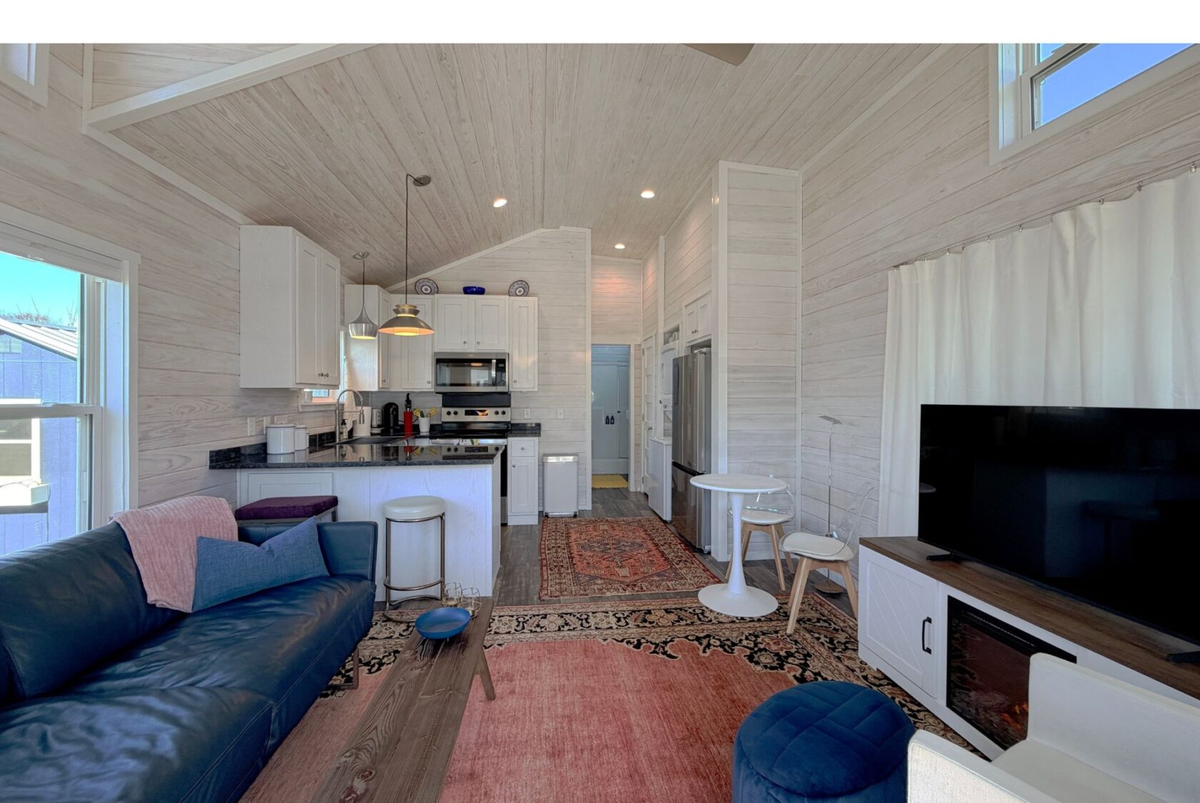 Bright, modern living room and kitchen at 49 Skipping Stone Lane with white walls, dark sofa, rugs, and a large TV.