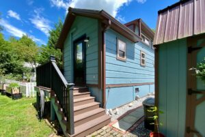Light blue tiny house on Just Wandering Lane, with brown trim, black door, and a small yard with greenery.