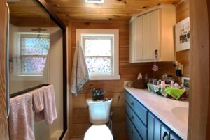 Small rustic bathroom on Just Wandering Lane with wood walls, blue vanity, shower, toilet, and frosted glass window.
