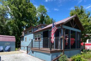 A small blue house with a porch and flag stands on Just Wandering Lane, nestled among trees on a sunny day.