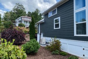 A modern house with dark siding on Just Wandering Lane, garden shrubs, and a hilltop home in the background.