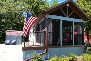 Charming small house on Just Wandering Lane with screened porch, shed, and an American flag in a sunny green yard.