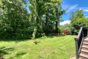 Backyard on Just Wandering Lane with green grass, tall trees, deck stairs, and a seating area with red chairs and umbrella.