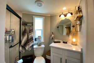 A modern bathroom on Just Wandering Lane with a shower, toilet, towels, and a round mirror above the white sink counter.