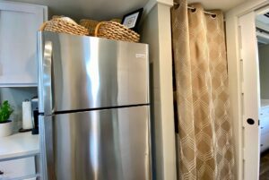 A stainless steel refrigerator on Just Wandering Lane, with baskets on top, by a beige curtain and white cabinets.