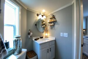 Small bathroom on Just Wandering Lane with a round mirror, white sink, shelves, and frosted window for natural light.