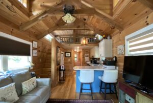 Cozy wooden cabin living area on Just Wandering Lane with sofa, TV, kitchen island, and lofted ceiling with exposed beams.