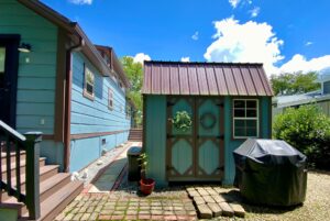 Charming blue shed with brown trim on Just Wandering Lane, beside a grill and blue house under a sunny sky.