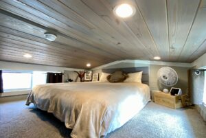 Cozy attic bedroom on Just Wandering Lane with sloped wooden ceiling, large bed, fan, and soft natural light.