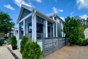 Modern small house with large windows on Just Wandering Lane, gray siding, fenced yard, and trees under a blue sky.