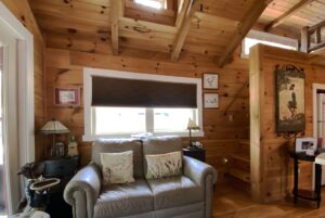 Cozy log cabin living room on Just Wandering Lane with a gray loveseat, pillows, and wood-paneled walls under a beamed ceiling.