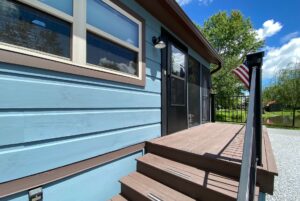 Small porch on Just Wandering Lane with brown steps, blue siding, railing, and flag beside a tree and pond.
