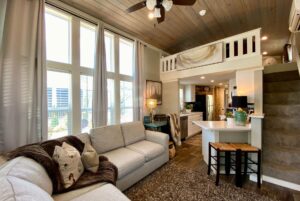 Cozy living room with large windows, loft, kitchen, and staircase in a modern tiny house on Just Wandering Lane.