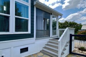 Outdoor steps on Just Wandering Lane lead to a screened porch by blue siding and white-framed windows under a sunny sky.