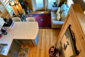A cozy living space on Just Wandering Lane with a small kitchen, sectional sofa, red rug, and wooden floors, viewed from above.