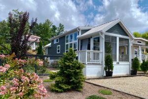 A modern tiny house on Just Wandering Lane with a screened porch, surrounded by gardens and trees under a partly cloudy sky.