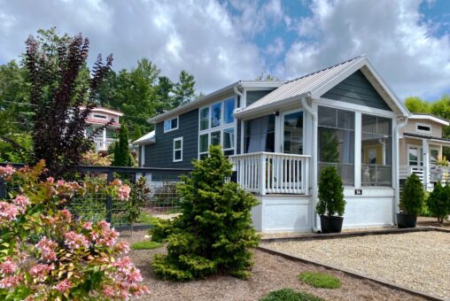A modern tiny house on Just Wandering Lane with a screened porch, surrounded by gardens and trees under a partly cloudy sky.
